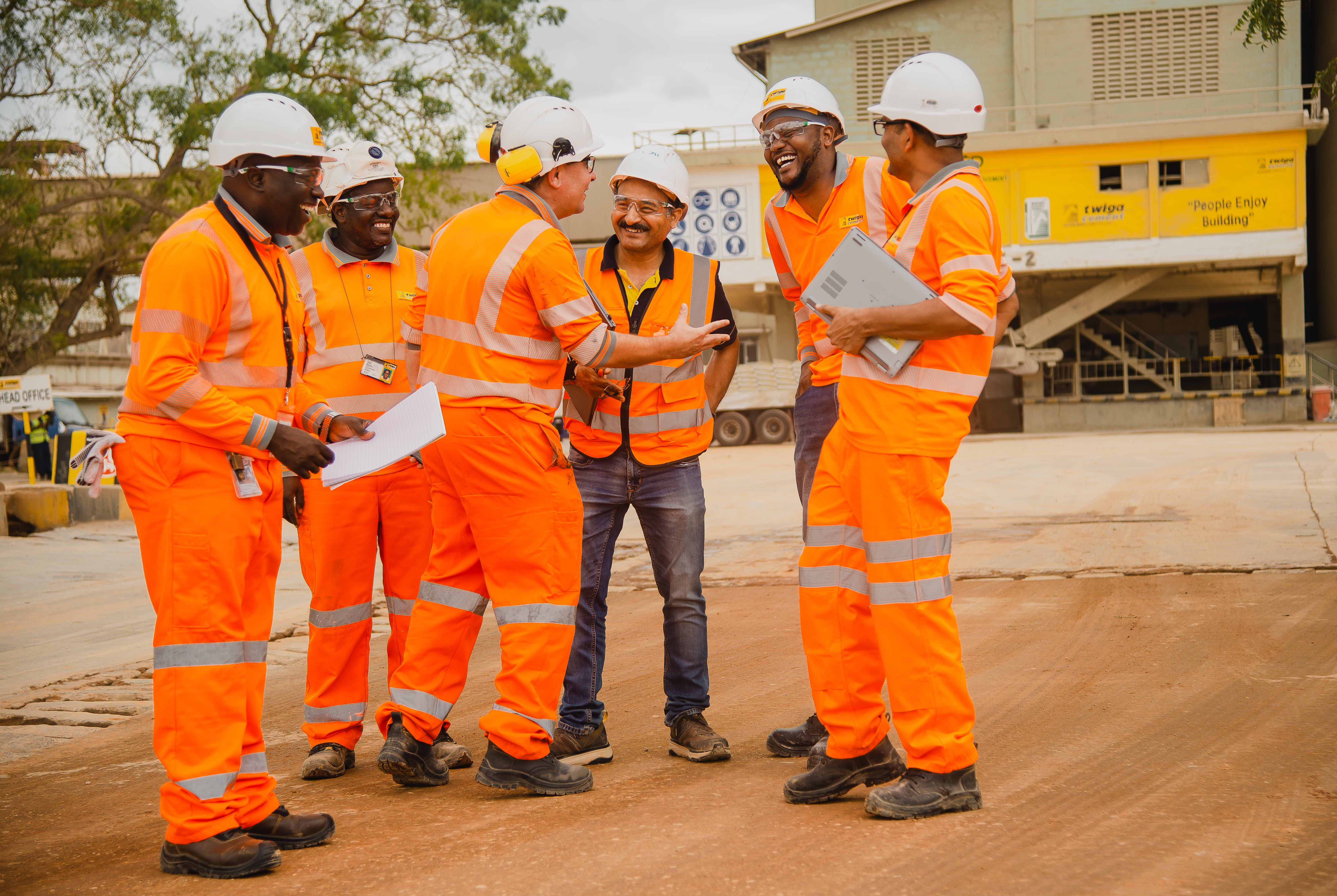  a group of six individuals wearing high-visibility orange safety vests and white hard hats. They are gathered in what appears to be a construction or industrial site. One person is holding open a set of plans or documents, which the group seems to be discussing. 
