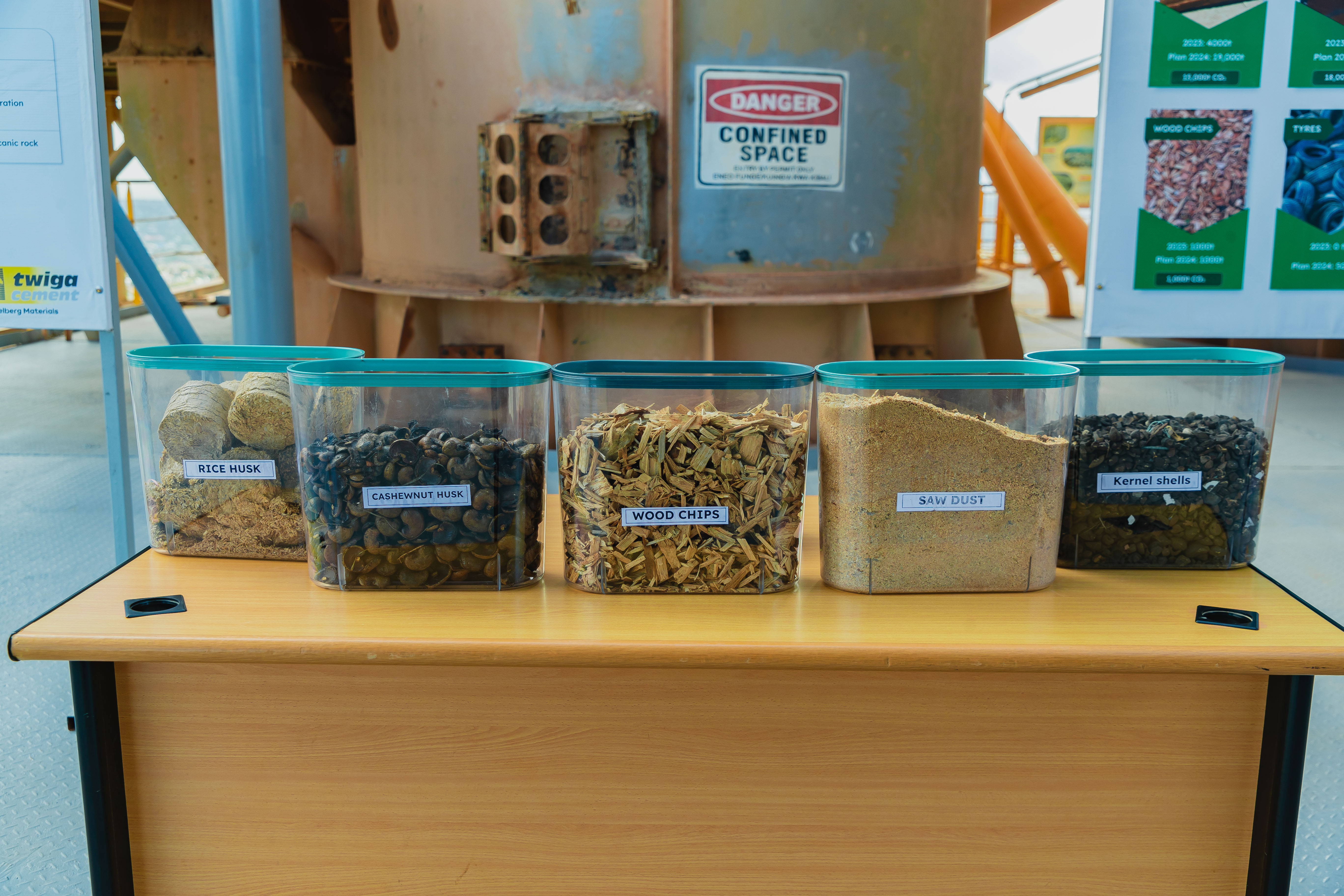five transparent containers, each filled with different materials labeled as “RICE HUSK,” “BAGASSE,” “WOOD CHIPS,” “SAW DUST,” and “KERNEL SHELLS.