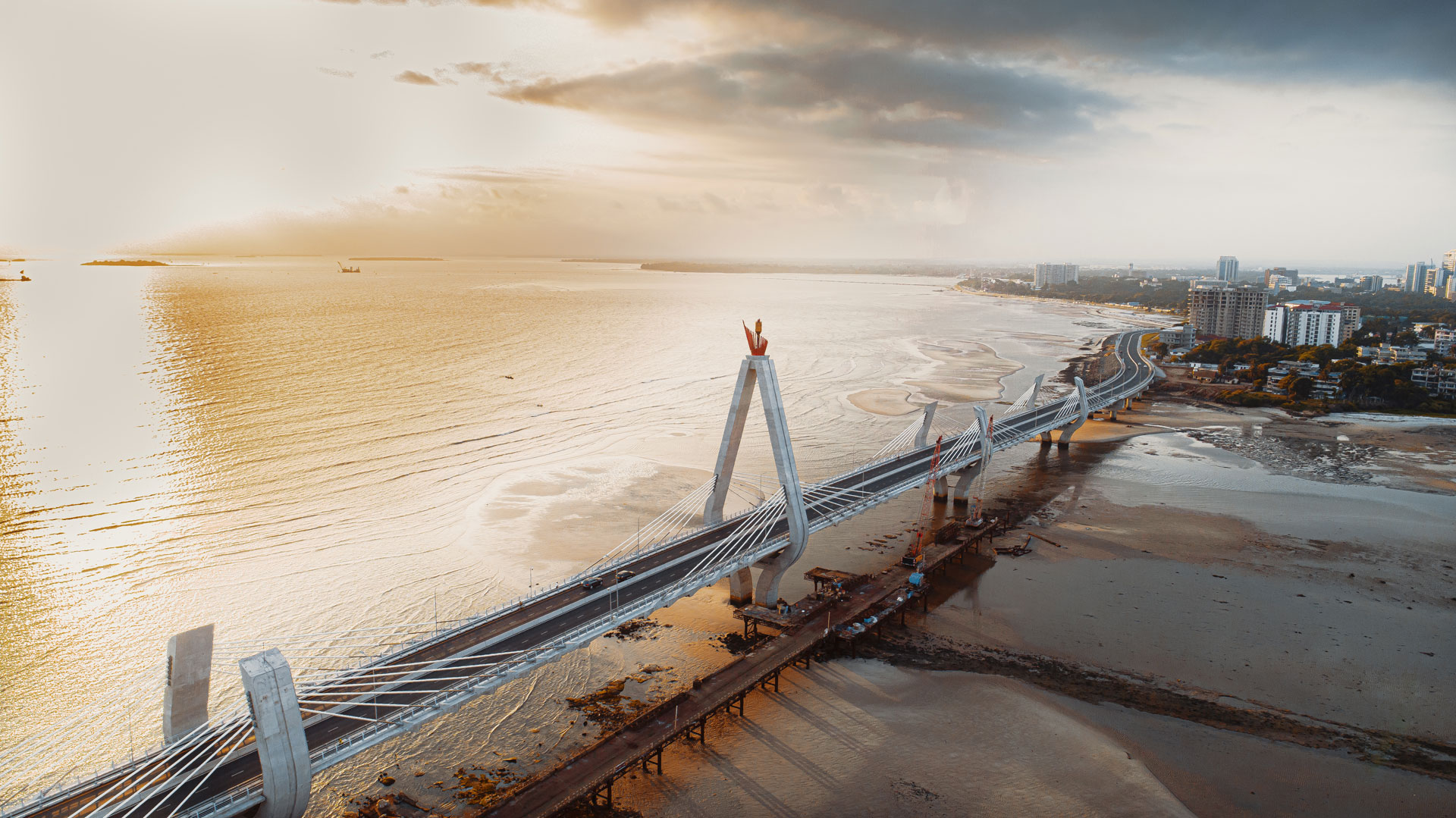 An aerial view of a large suspension bridge extending over a body of water, with a city skyline visible in the distance. The bridge features distinctive towers and cables, and the lighting suggests it is either sunrise or sunset, casting a warm glow over the scene