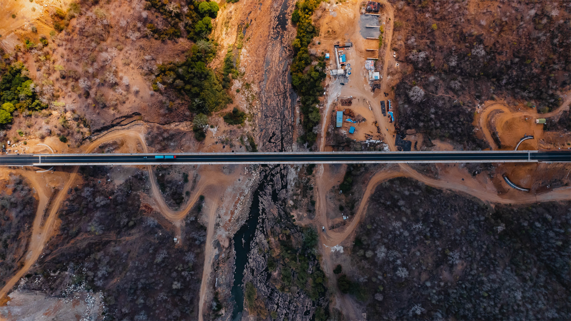 An aerial view of a bridge spanning a rugged, arid canyon. Below the bridge, dirt paths and a parking area with several vehicles are visible. The terrain is sparse with vegetation, highlighting the bridge's role in connecting remote areas