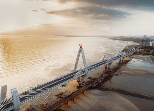 An aerial view of a large suspension bridge extending over a body of water, with a city skyline visible in the distance. The bridge features distinctive towers and cables, and the lighting suggests it is either sunrise or sunset, casting a warm glow over the scene